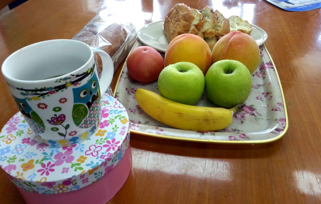 productivity hack treats image of wood table that has a tray of fruit including three peaches, two green apples, and a banana. Behind the tray is a place of sliced pastry bread. In the front to the left is a coffee mug with an owl design.